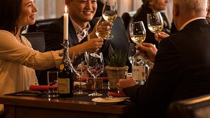 Un grupo de personas brindando con copas de vino en una mesa con una vela, botellas de vino y una planta en maceta.