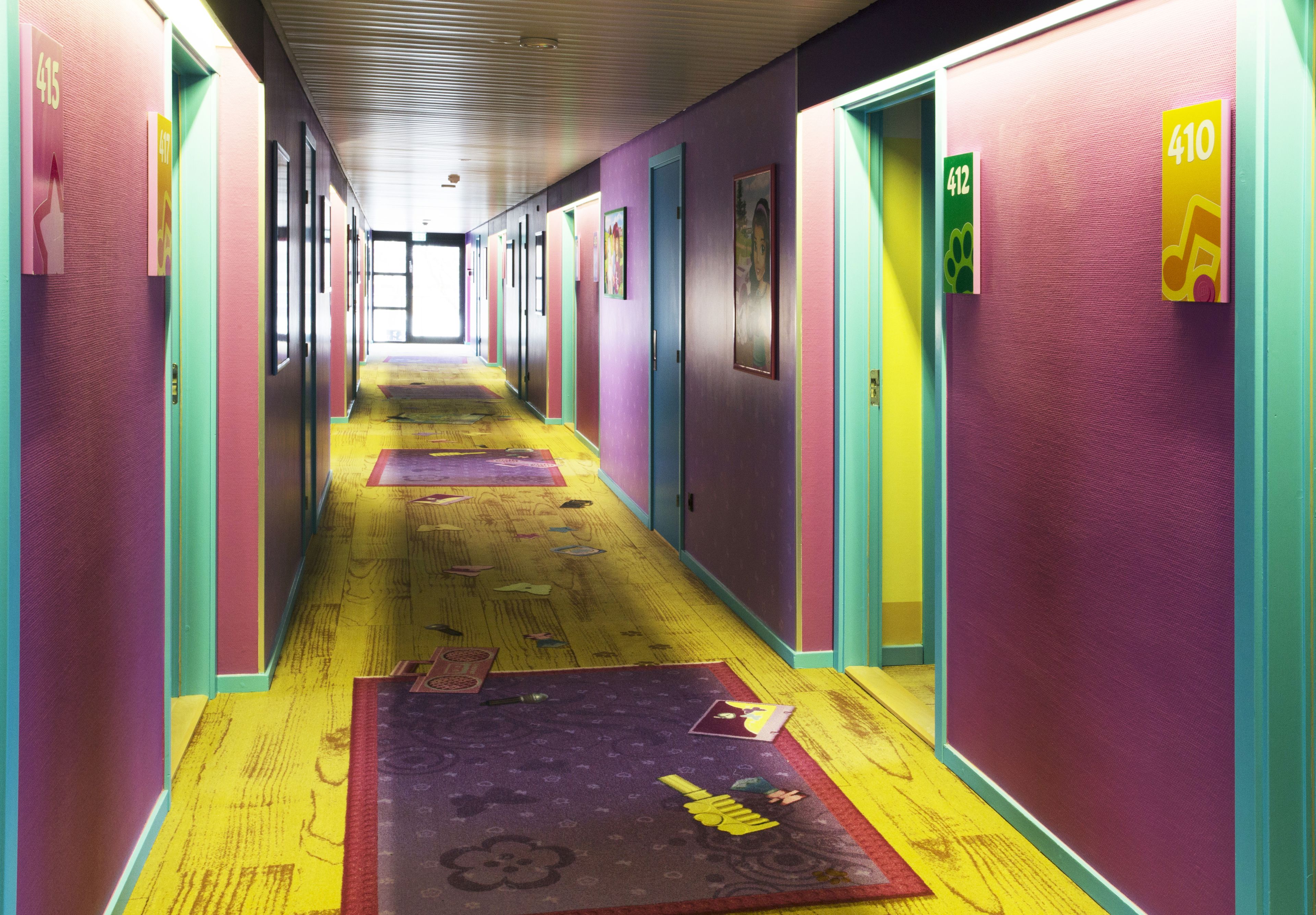 Un pasillo luminoso con puertas numeradas, suelo de madera y alfombras estampadas.
