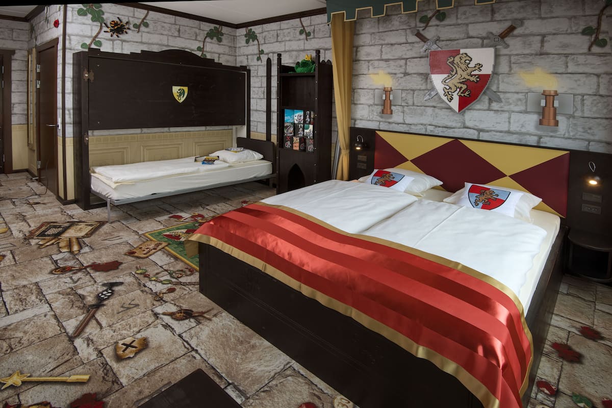 A bedroom with a large bed, a smaller bed, and a wardrobe. There are decorative elements on the walls and a carpet with a medieval theme.