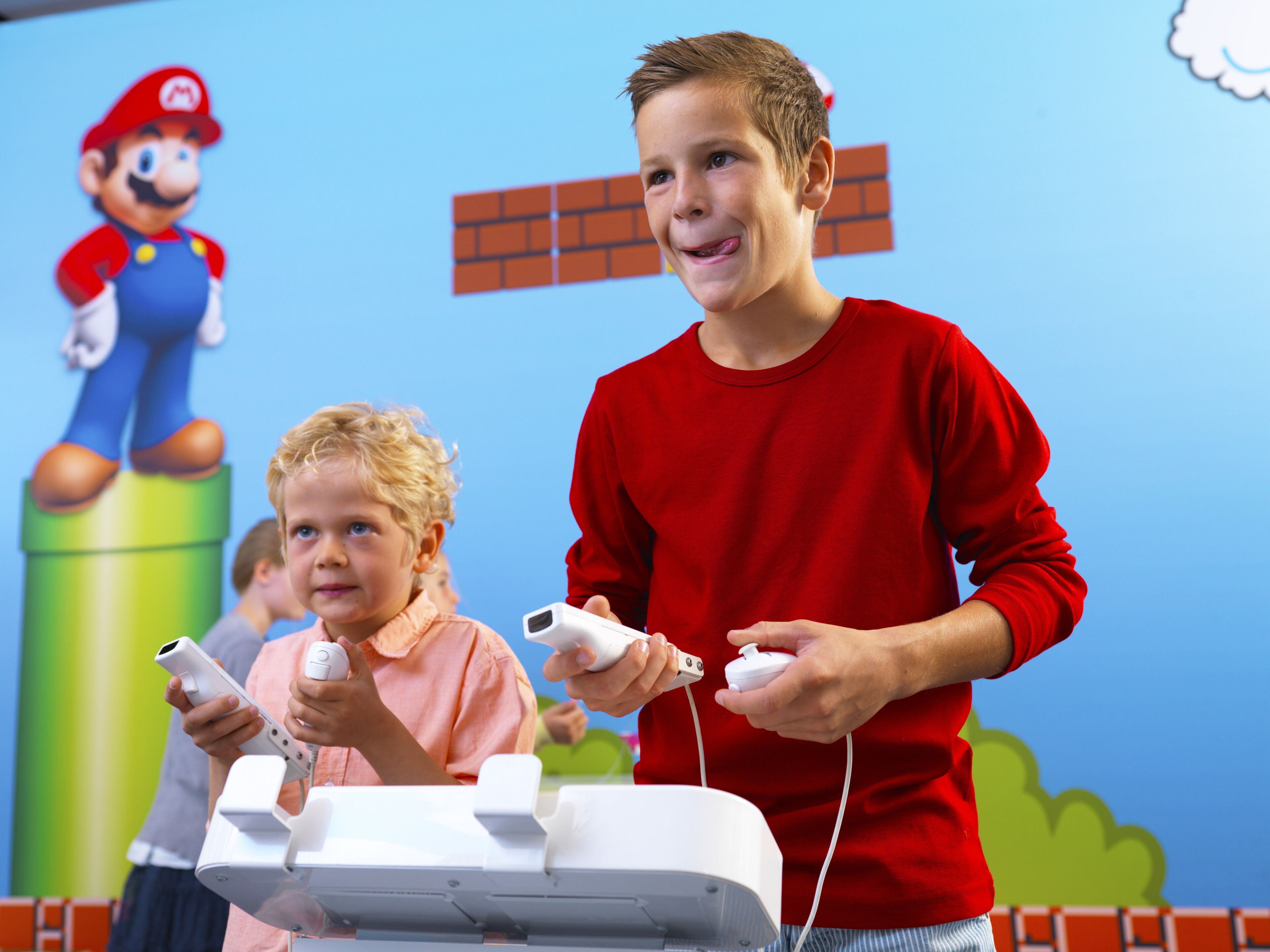 A child and an adult playing a video game with a Mario character in the background.