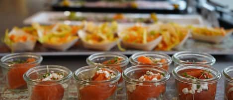 Una bandeja con alimentos, incluyendo pequeños vasos con salmón ahumado y adornos, y otra bandeja más grande con ensaladas variadas.