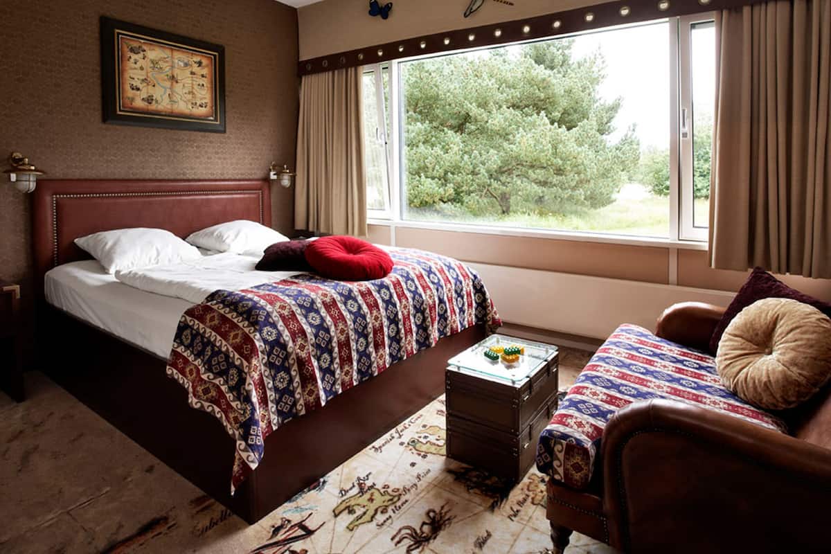 A hotel room with a large bed, a wooden headboard, a nightstand with a lamp, a window with curtains, and a view of greenery outside.