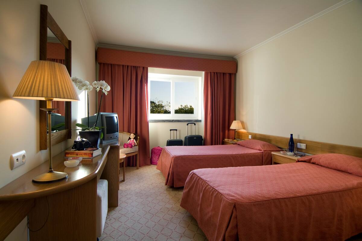 Habitación de hotel con dos camas, un escritorio con lámpara, televisión y ventana con cortinas, y vista a zonas verdes.