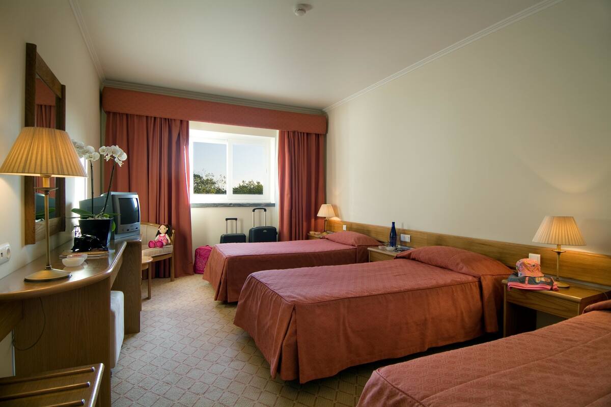 Habitación de hotel con dos camas, un escritorio, una silla, un televisor, una lámpara y una ventana con cortinas.