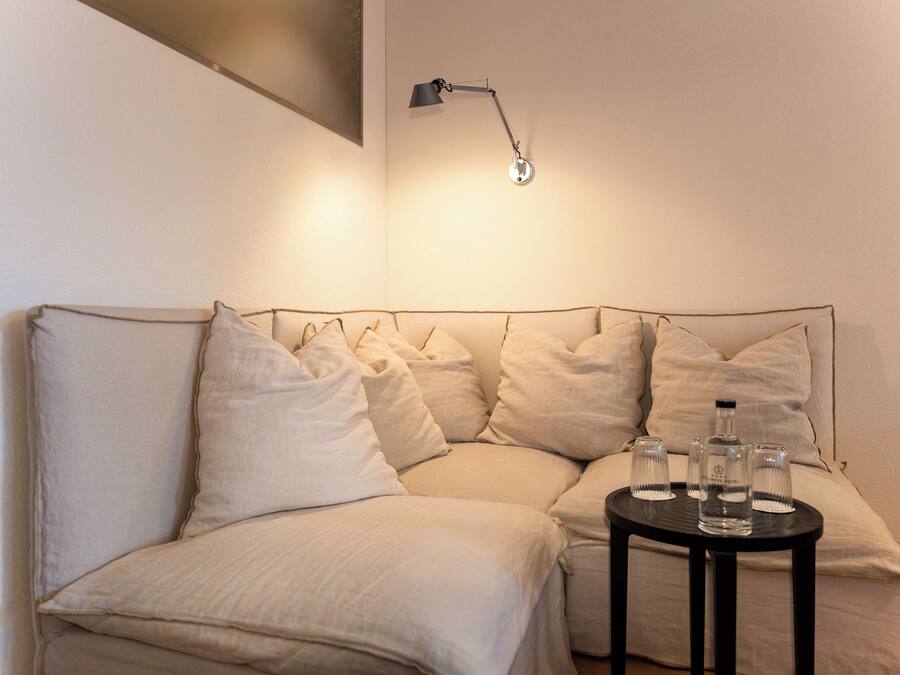 Ein Sofa mit mehreren Kissen, ein kleiner Tisch mit Gläsern und einer Flasche sowie eine Wandleuchte.