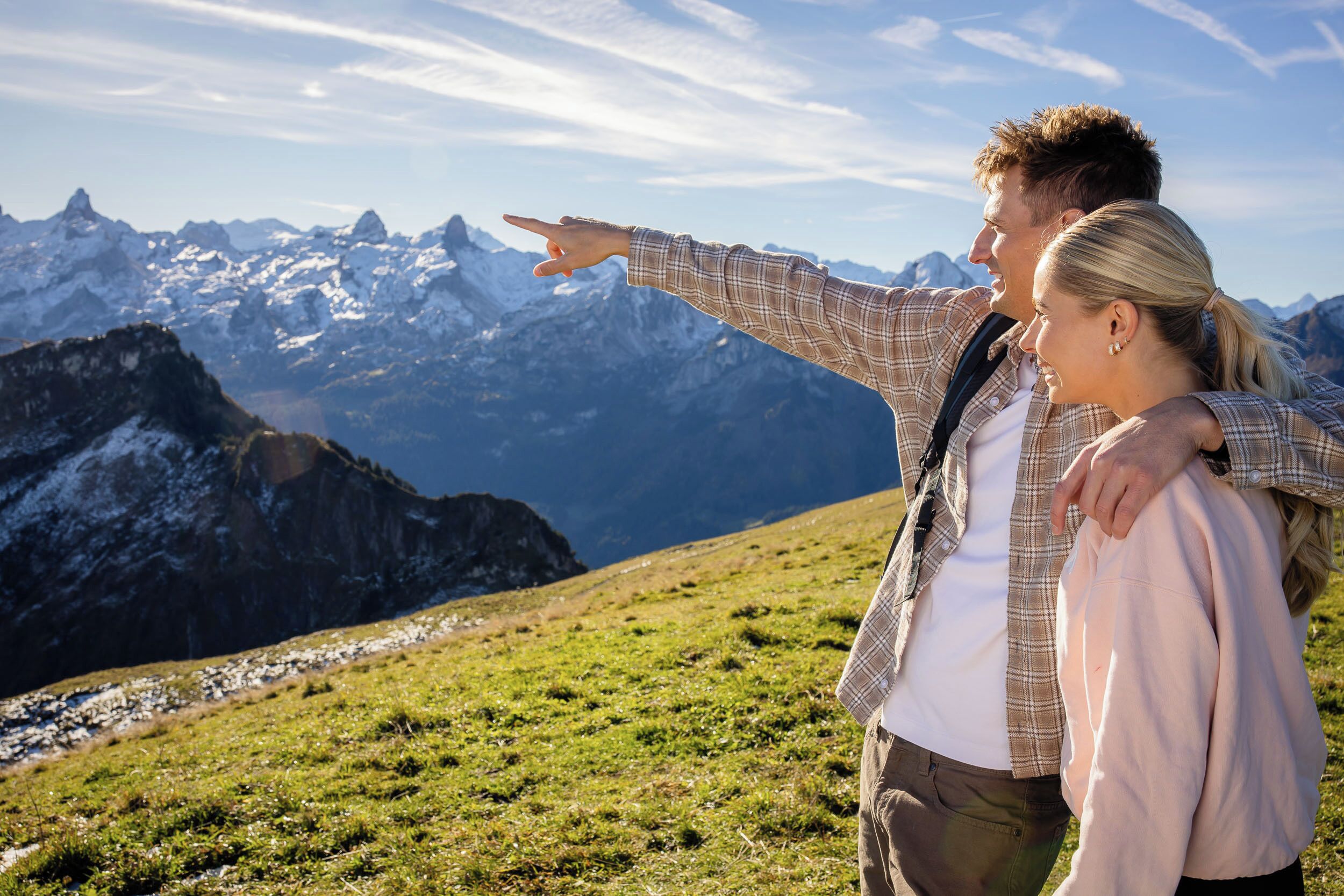 Un couple se tient sur une colline herbeuse, avec un paysage montagneux en arrière-plan.