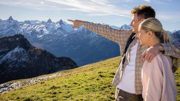 Un couple se tient sur une colline herbeuse, avec un paysage montagneux en arrière-plan.
