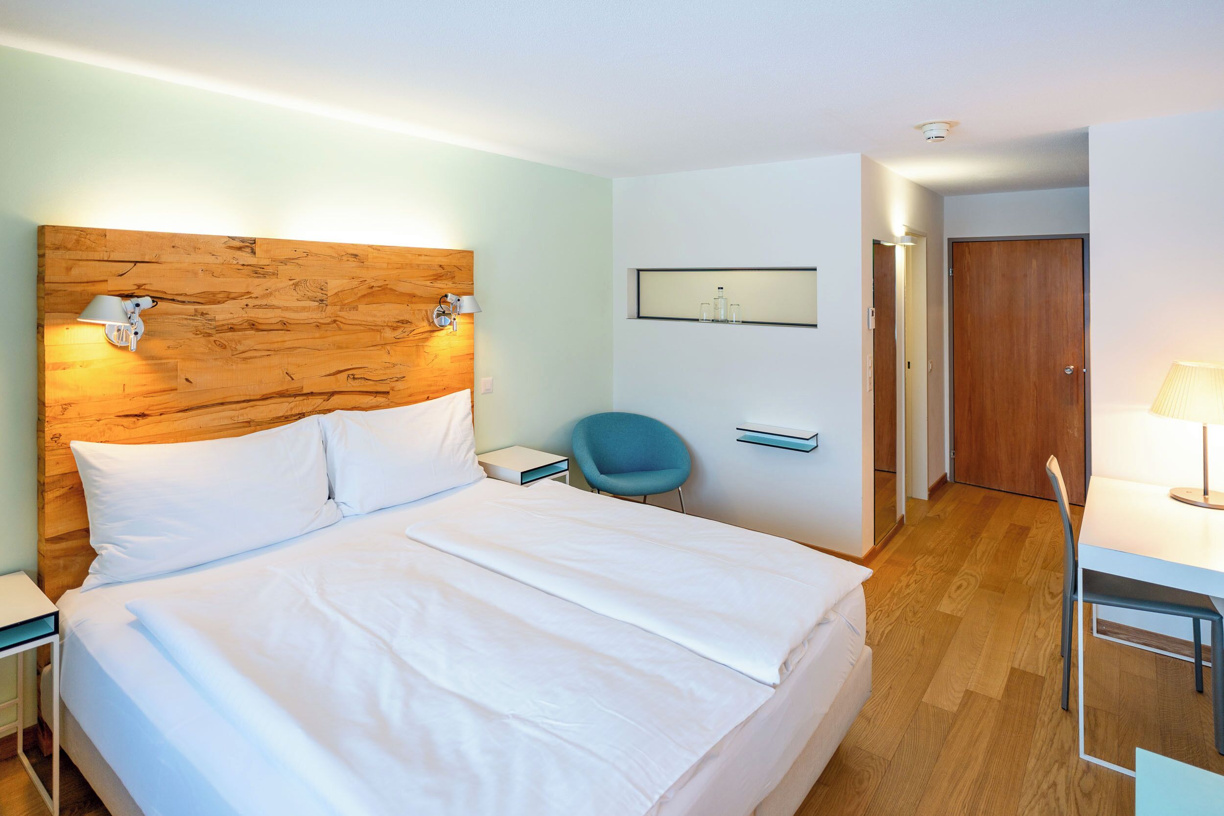 A hotel room with a large bed, a nightstand, a desk, a chair, and a wooden headboard.