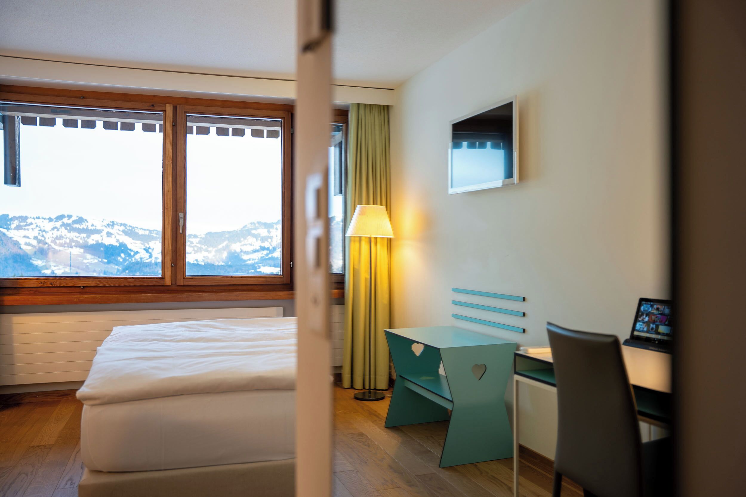Et hotelværelse med seng, skrivebord, stol, lampe og et vindue med udsigt til bjerge.