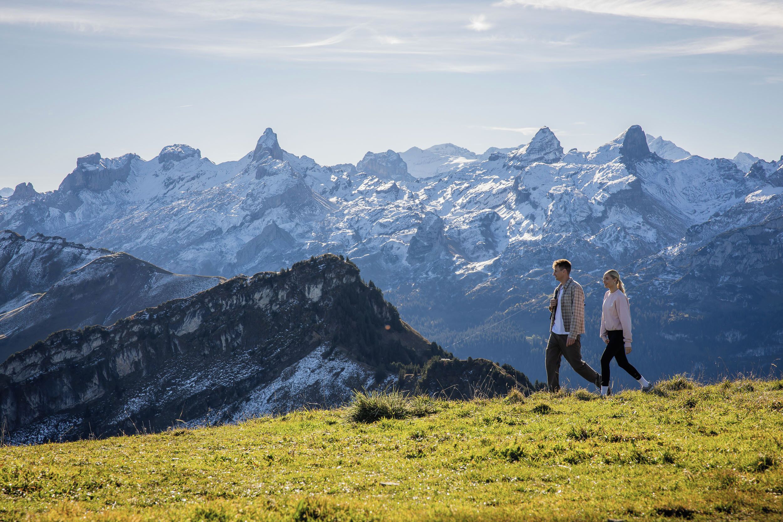 A couple walking on a grassy mountain path with snow-capped peaks in the background.