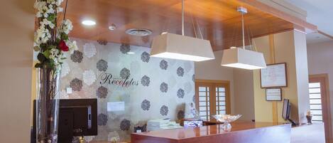 A reception desk with a wooden finish, a floral wallpaper background, and a large vase with flowers.
