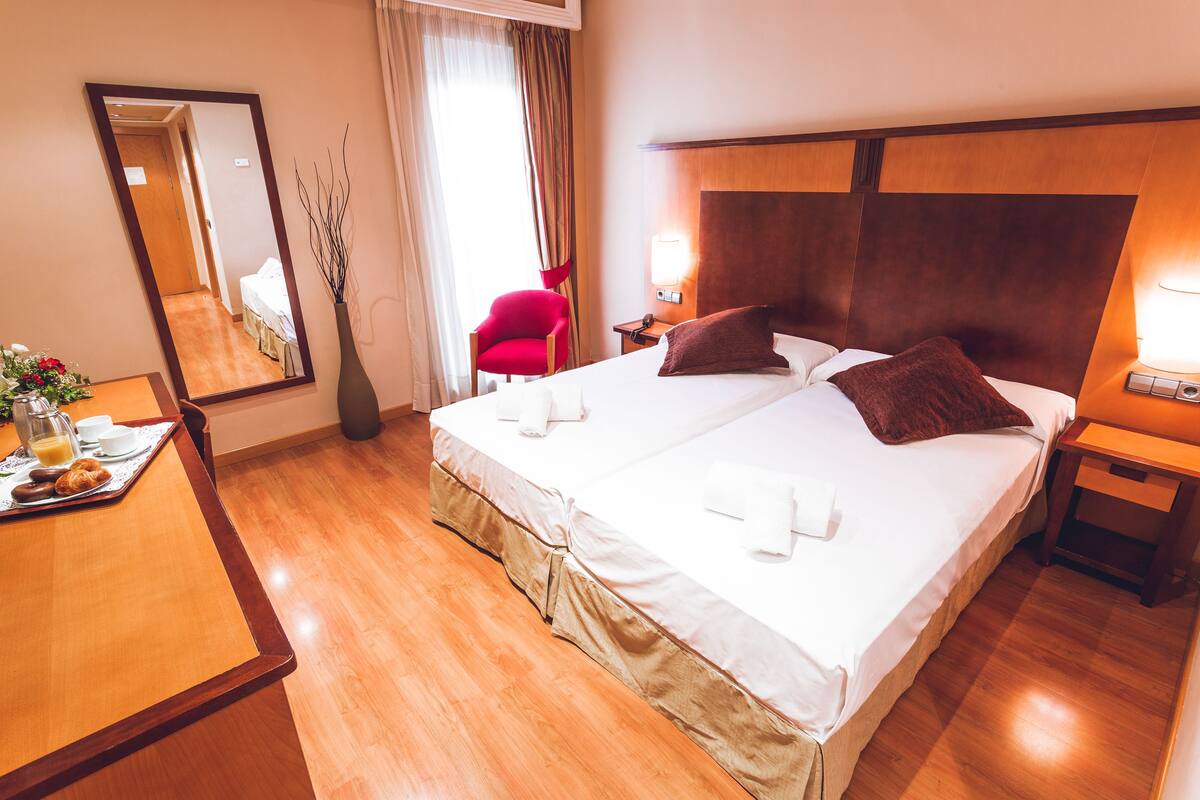A hotel room with a large bed, a wooden headboard, a bedside table with a lamp, a red chair, and a mirror on the wall.
