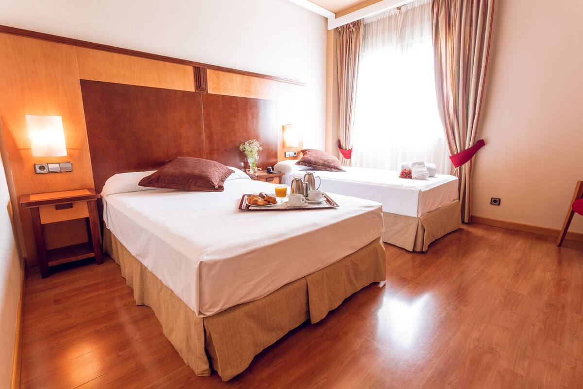 A hotel room with two beds, wooden headboards, a nightstand, a tray with breakfast items, and a window with curtains.