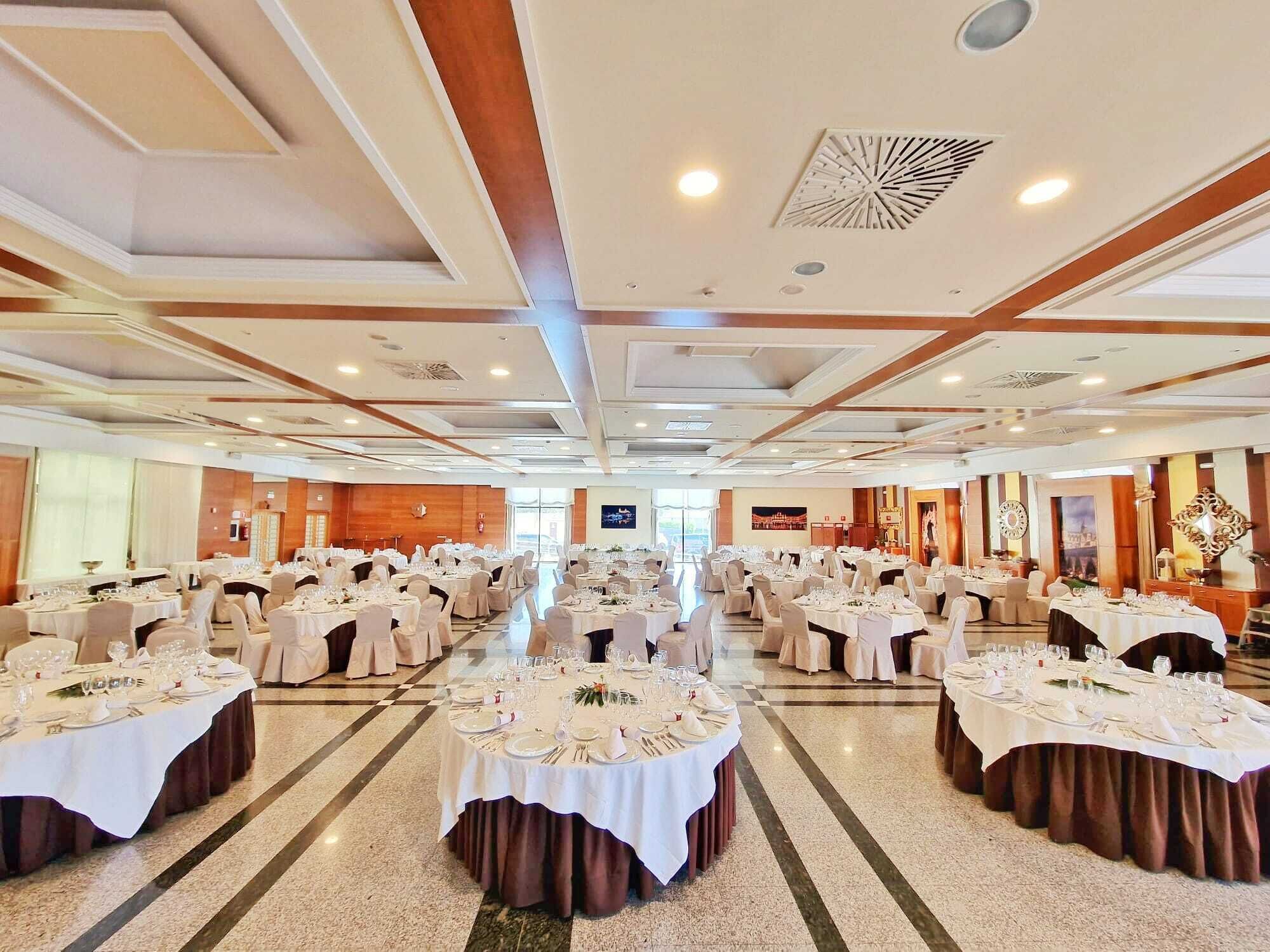 A large banquet hall with multiple round tables set for dining, featuring white tablecloths and formal table settings.