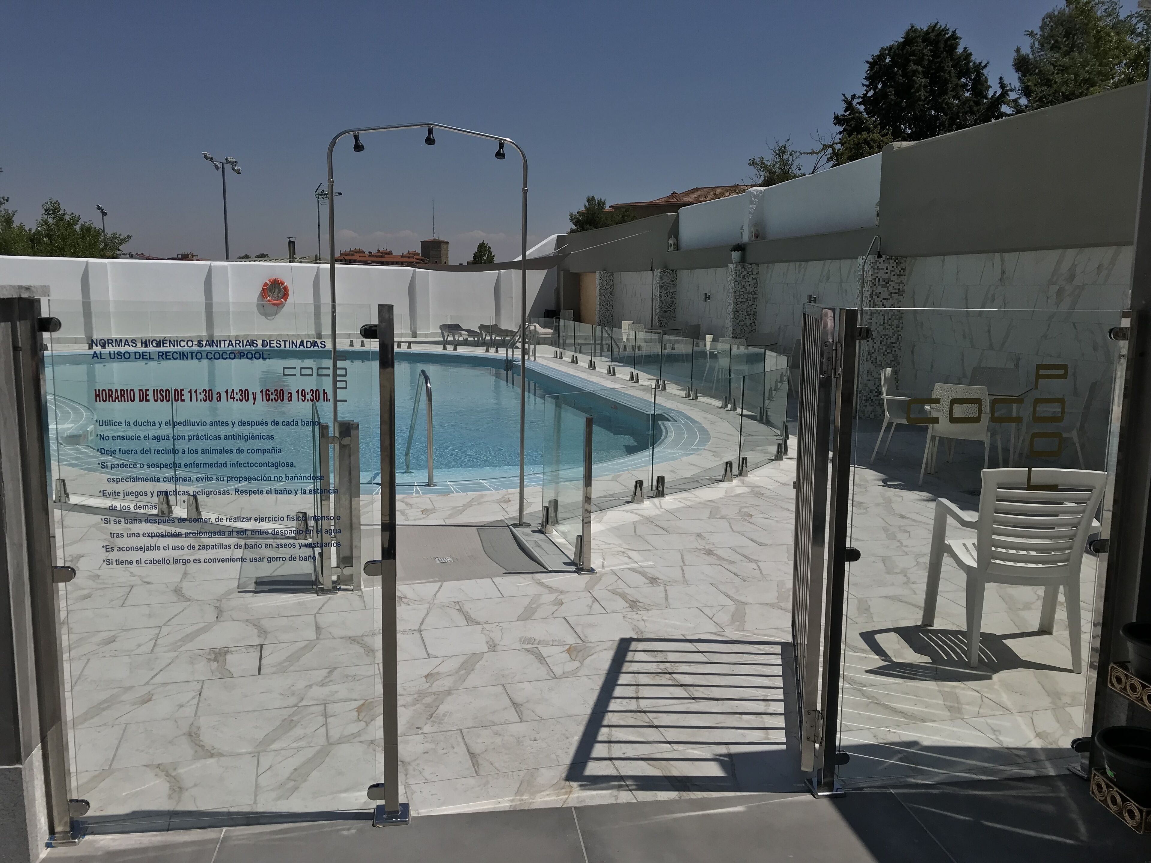 Piscina con recinto de vidrio, rodeada por un patio empinado con sillas y puerta de vidrio.
