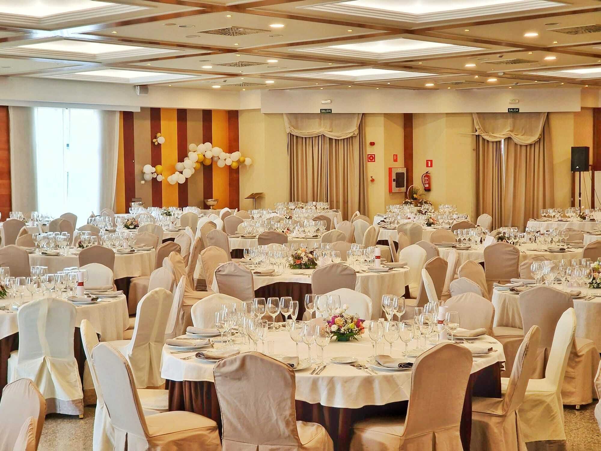 Un salón de banquetes con mesas redondas dispuestas para comer, sillas blancas e iluminación empotrada en el techo.