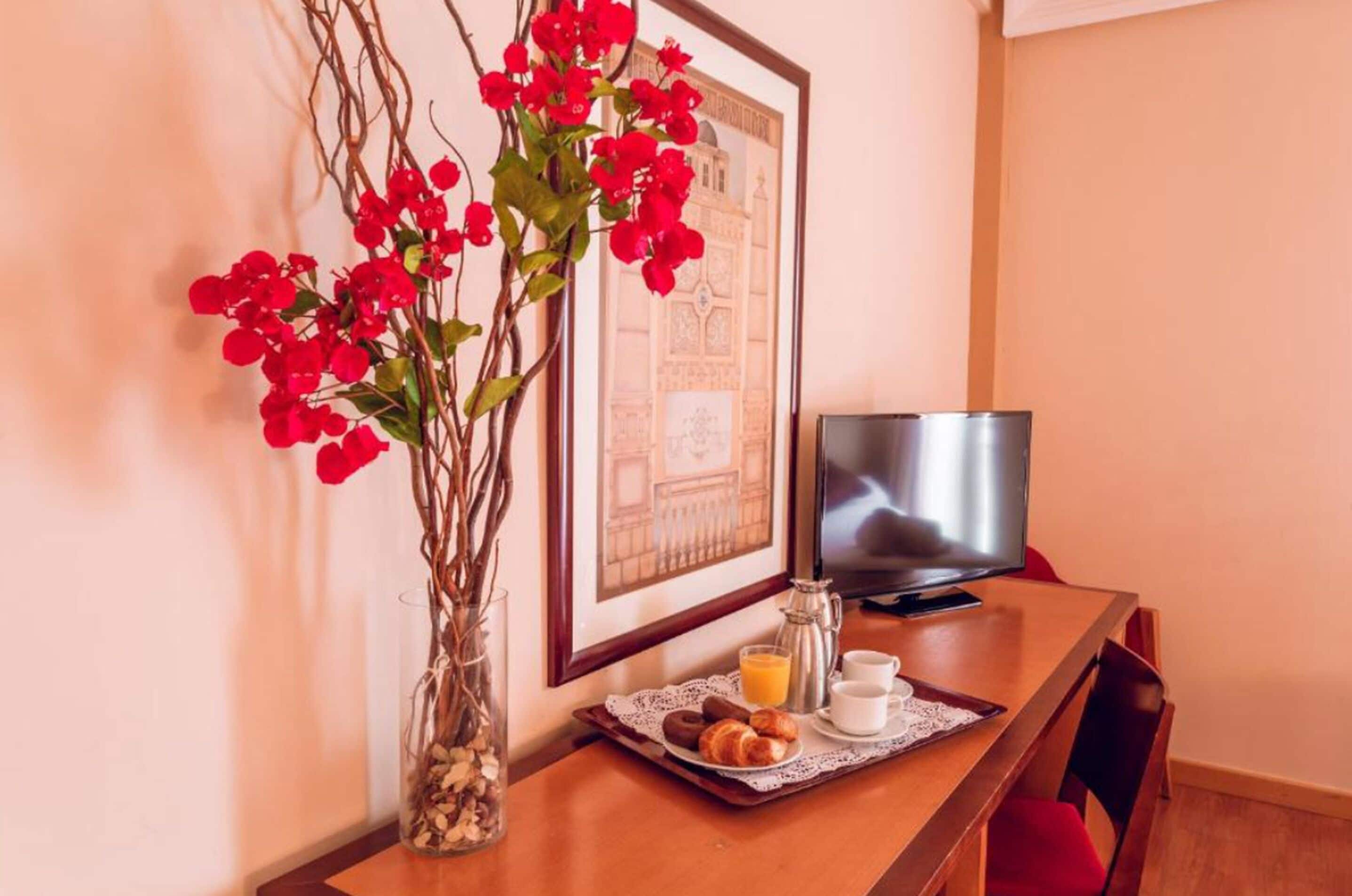 A breakfast setup with a tray of croissants, juice, and coffee, a vase with red flowers, and a framed picture on a wooden table.