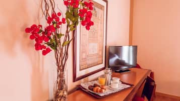 A breakfast setup with a tray of croissants, juice, and coffee, a vase with red flowers, and a framed picture on a wooden table.
