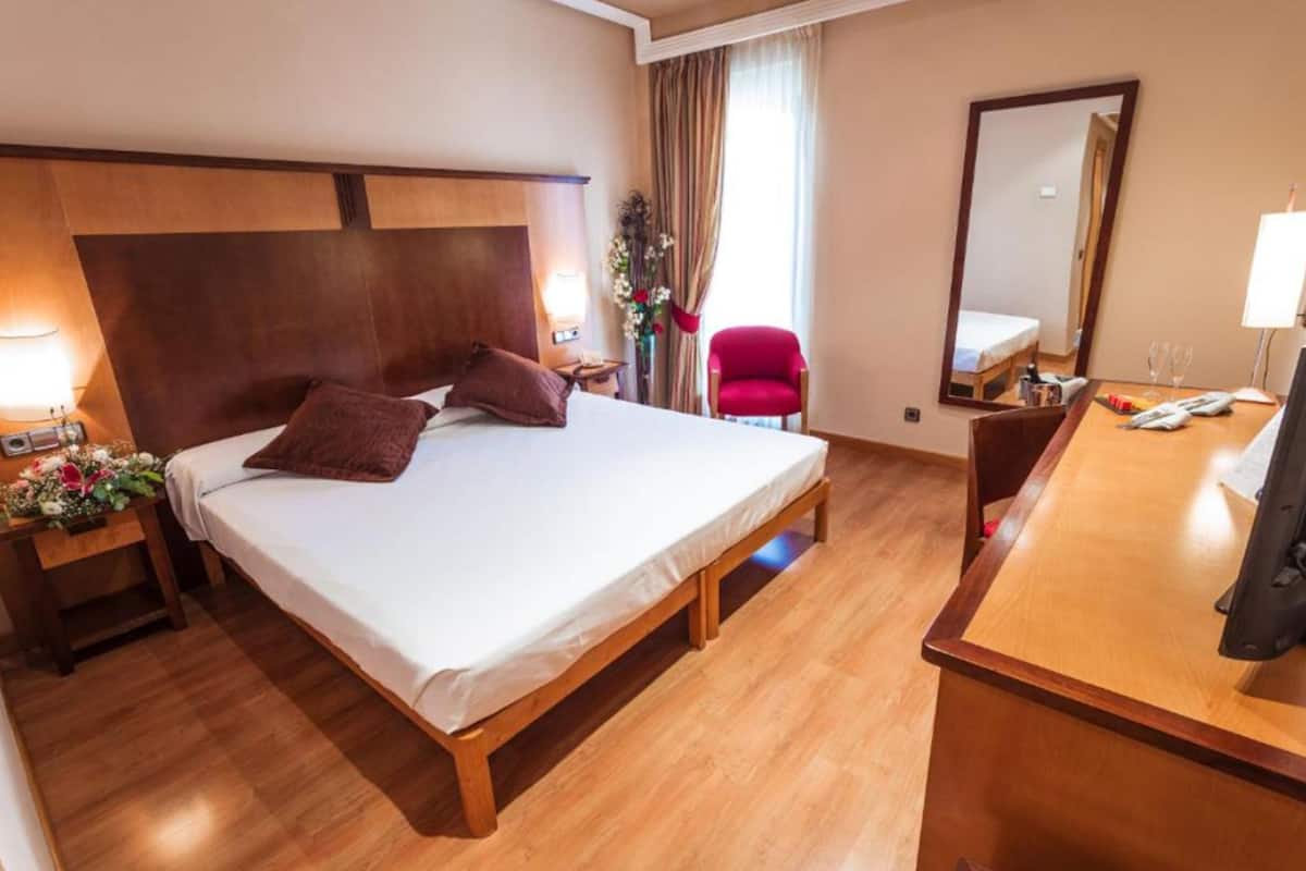 A hotel room with a large bed, wooden headboard, a desk with a chair, a mirror, and a small table with a lamp.