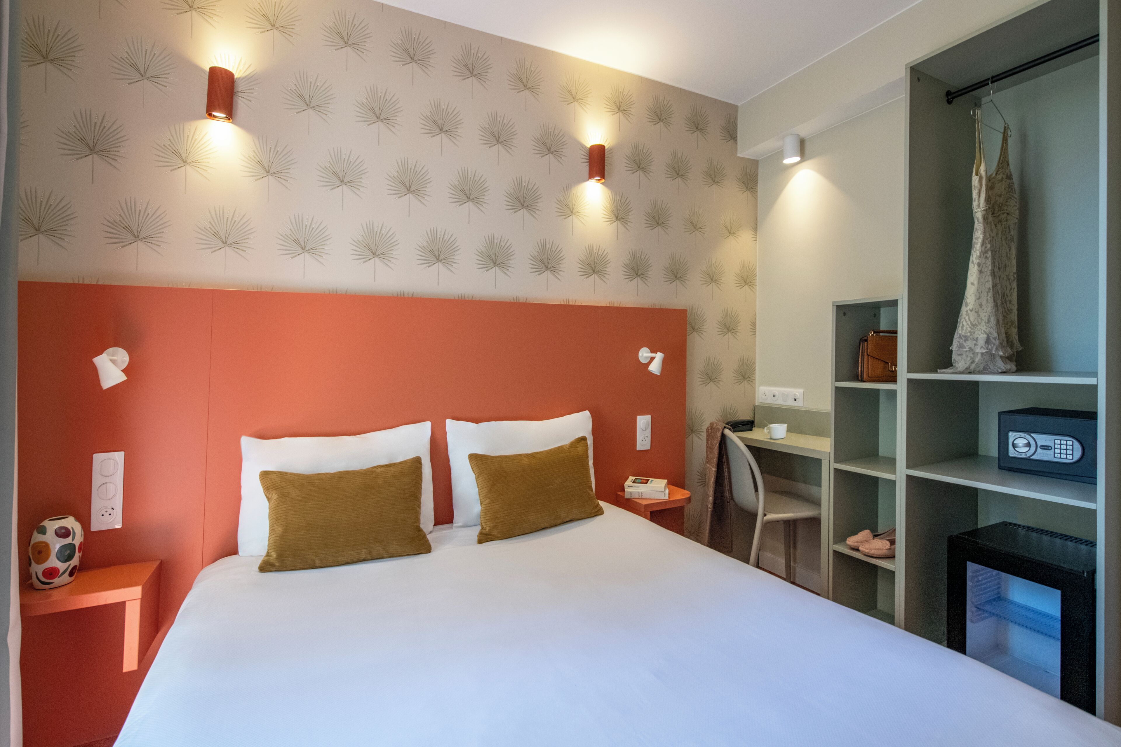 Une chambre d’hôtel avec un grand lit, deux lampes fixées au mur, un petit bureau et une étagère sur laquelle est posé un objet décoratif.