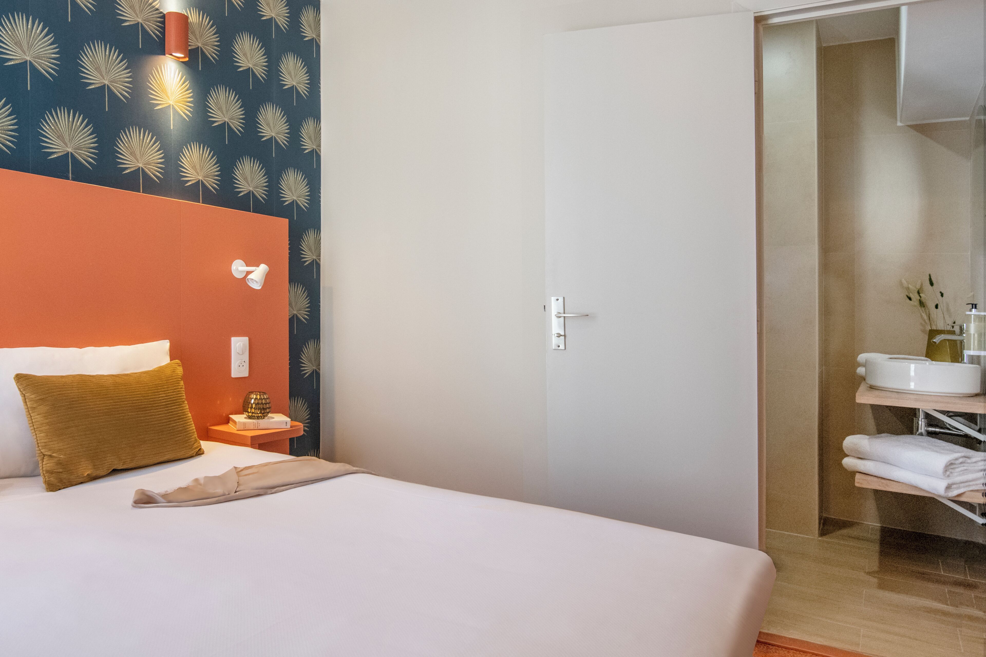 Een hotelkamer met een bed, een nachtkastje, een wandlamp, een deur en een zichtbare badkamer via een geopende deur.