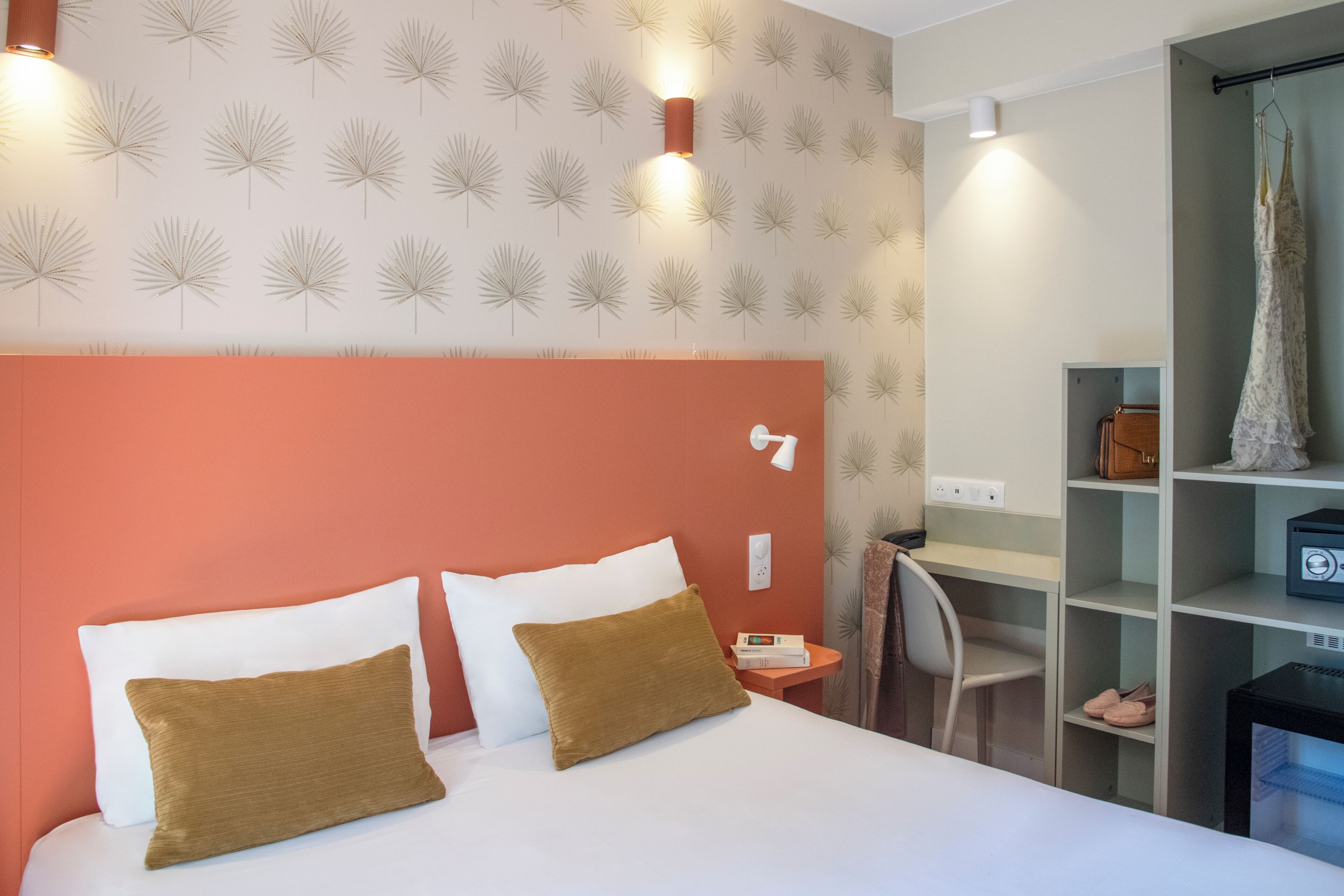 Hotelkamer met een bed, kussens, een nachtkastje, een stoel, een bureau, een plank met spullen en een muur met een bladmotief.