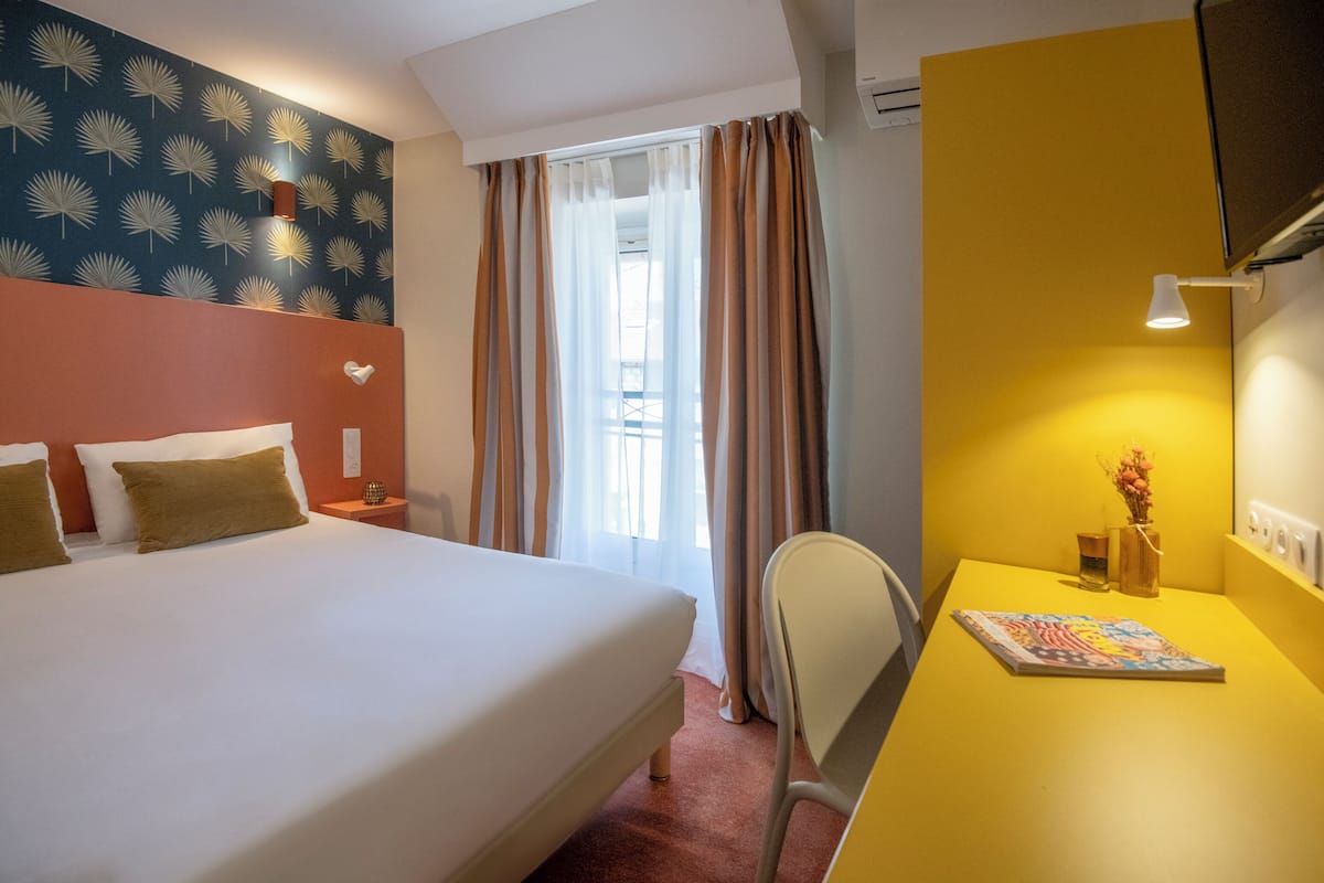 A hotel room with a bed, a desk, a chair, a window with curtains, a wall with a leaf pattern, and a wall-mounted lamp.