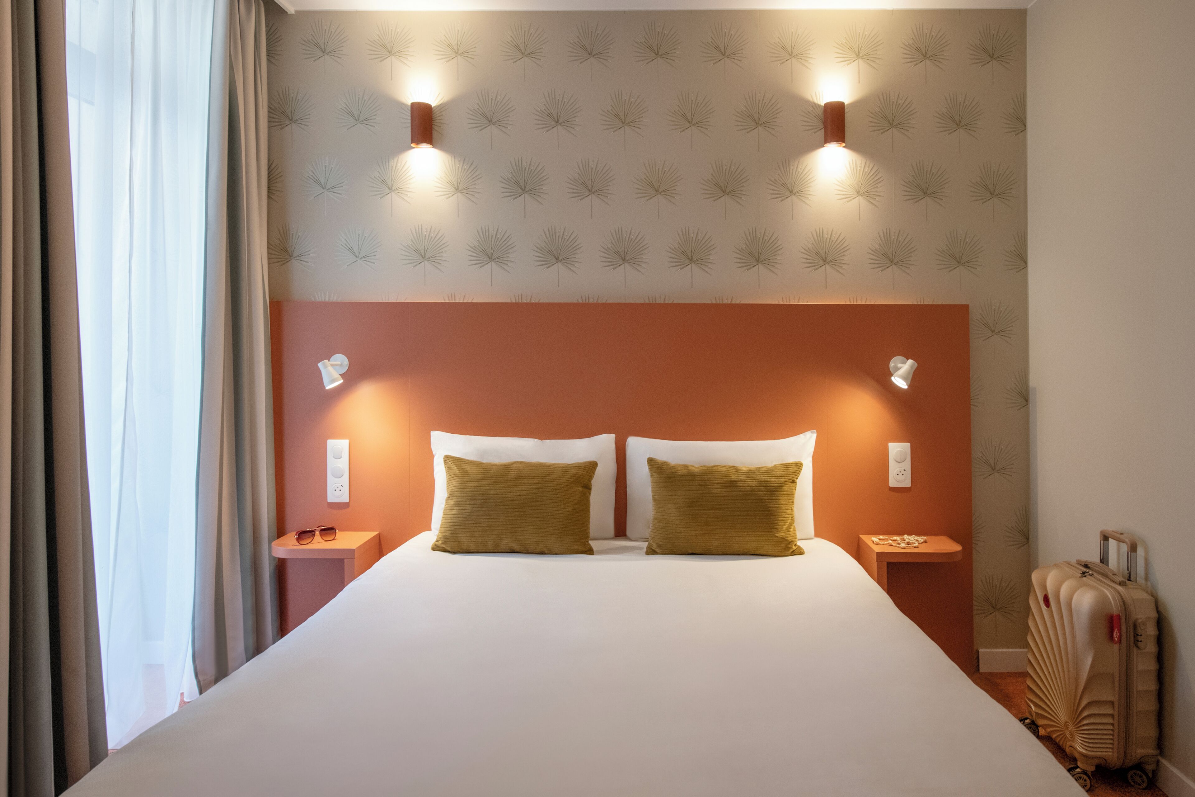 A hotel room with a large bed, two bedside lamps, a nightstand, a suitcase, and a window with curtains.