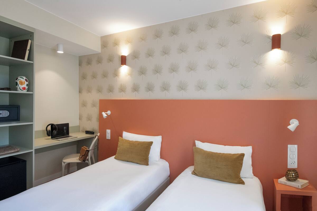 A hotel room with two beds, a desk with a laptop and headphones, a bookshelf, and wall-mounted lighting.
