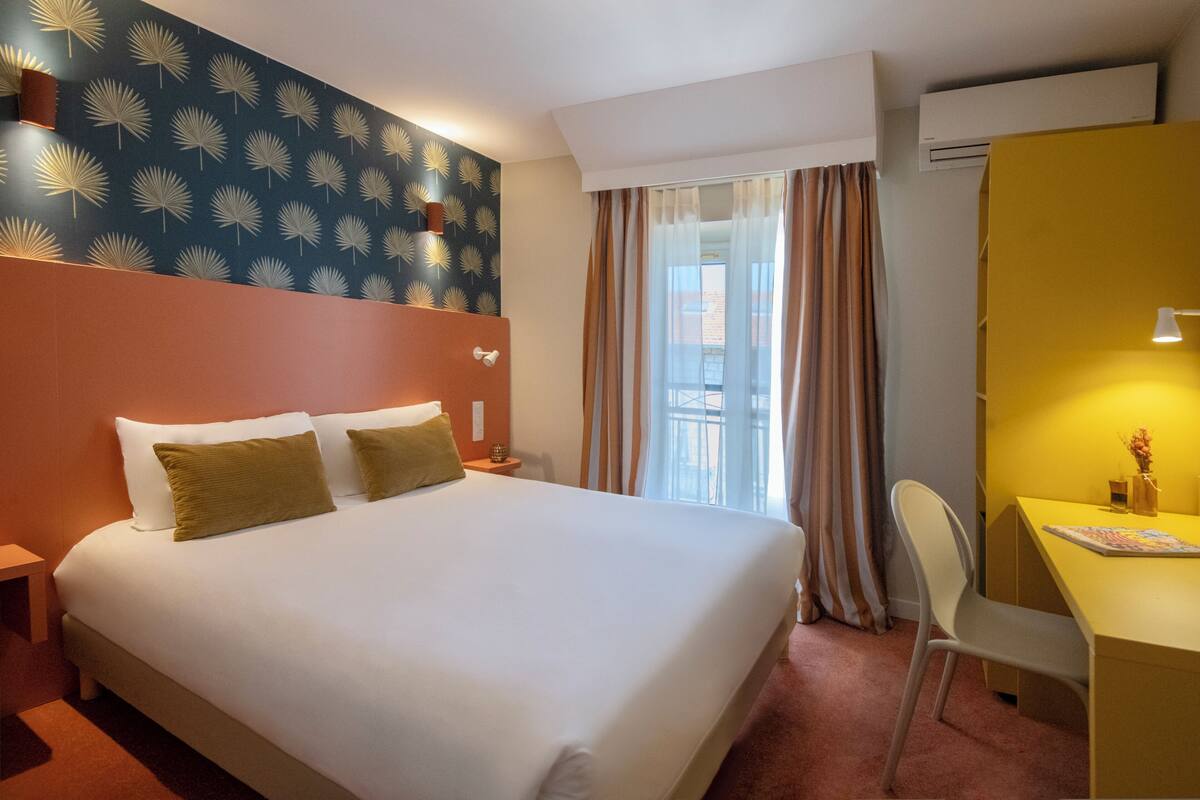 A hotel room with a large bed, a desk, a chair, a nightstand, and a window with curtains.