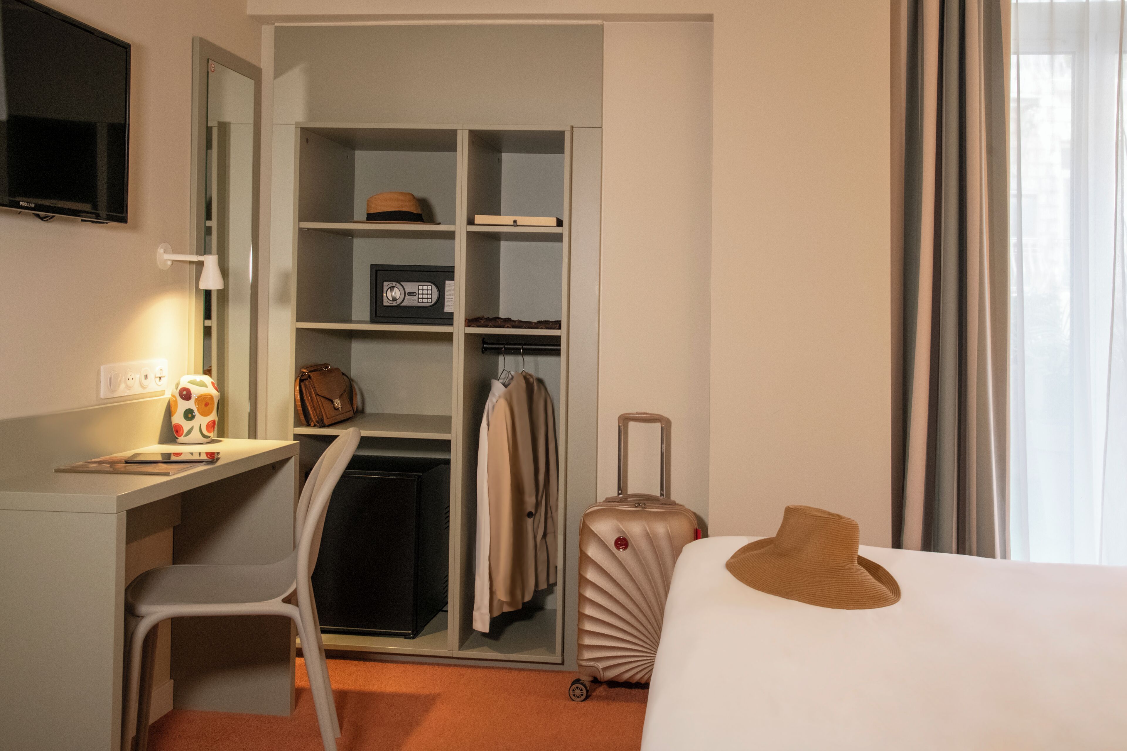 Una habitación de hotel con cama, un escritorio con lámpara, un armario y una maleta.