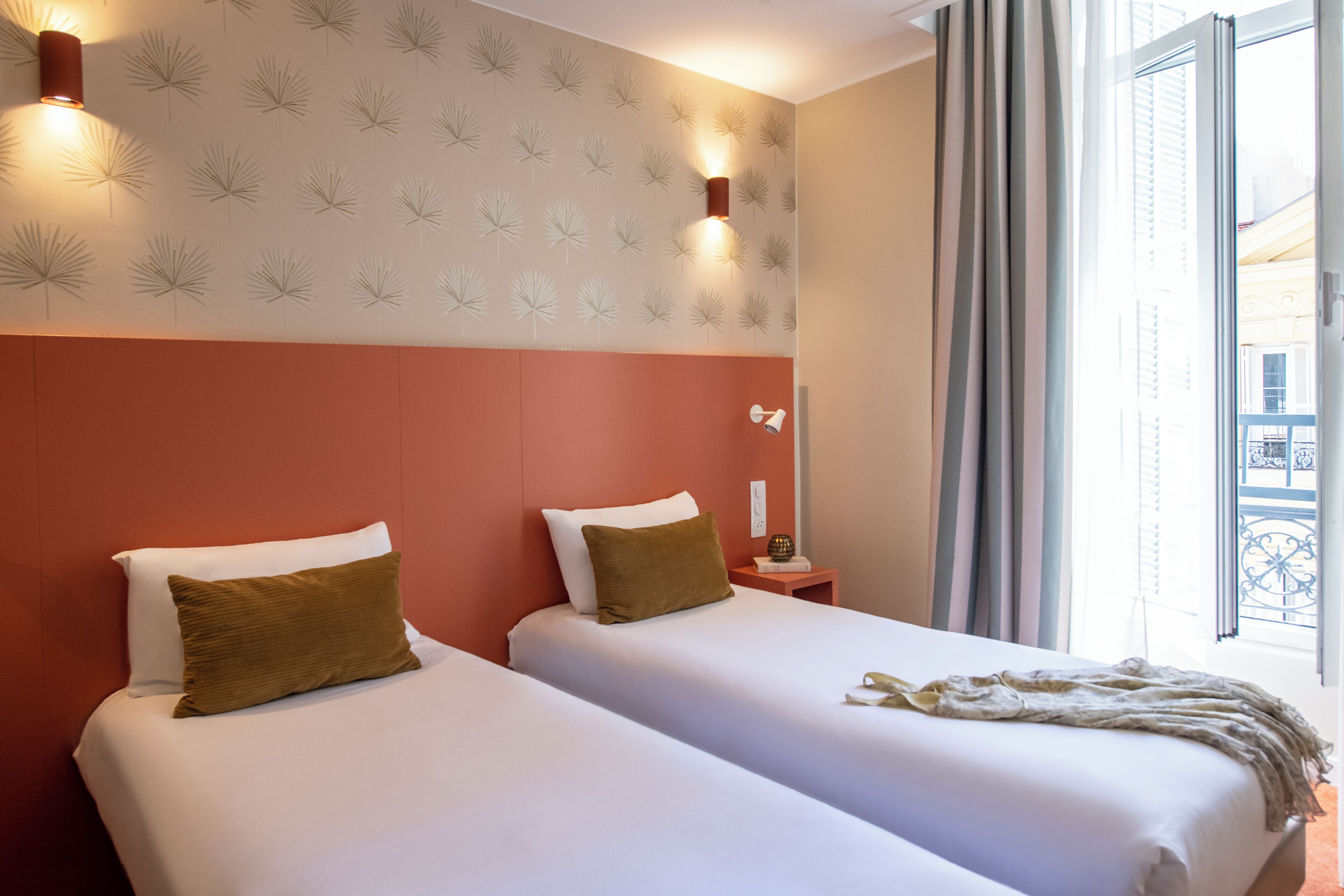 A hotel room with two beds, a patterned wall, a window with curtains, and a nightstand with a lamp.