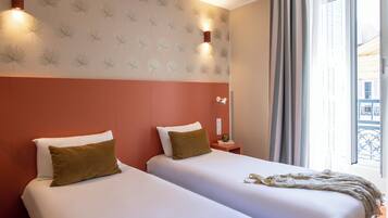 A hotel room with two beds, a patterned wall, a window with curtains, and a nightstand with a lamp.