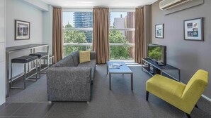 Un salon moderne avec un canapé gris, un fauteuil jaune et un téléviseur posé sur un meuble. De grandes fenêtres sont ornées de rideaux, un climatiseur est installé et des tableaux encadrés décorent les murs.