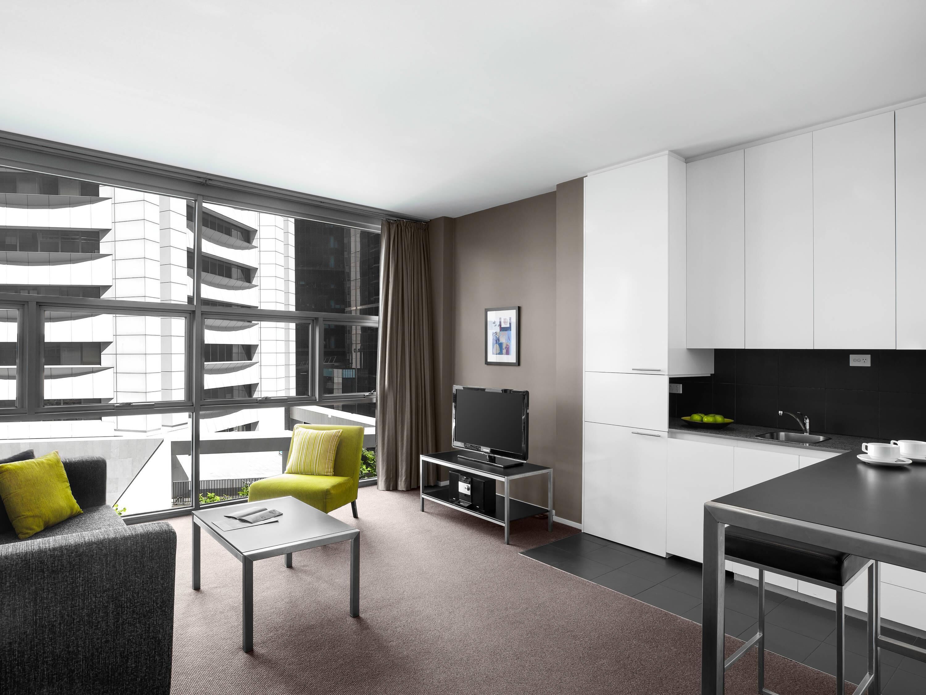 A modern hotel room with a sofa, a small table, a TV, a kitchen area with a sink and cabinets, and large windows offering a view of a building.
