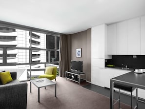 A modern hotel room with a sofa, a small table, a TV, a kitchen area with a sink and cabinets, and large windows offering a view of a building. - Exhibition Street Apartments by CLLIX (Melbourne)