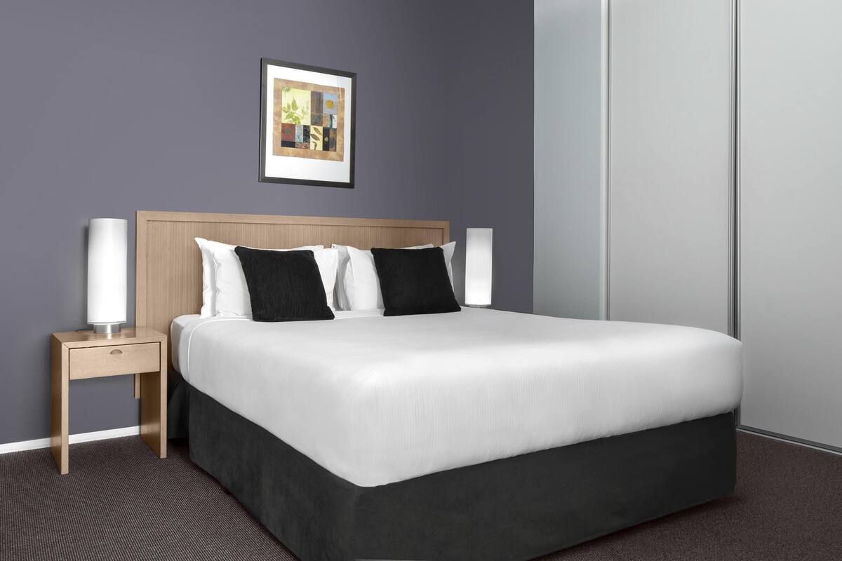 A neatly made bed with white and black bedding, a wooden headboard, a bedside table with a lamp, and a framed picture on the wall.