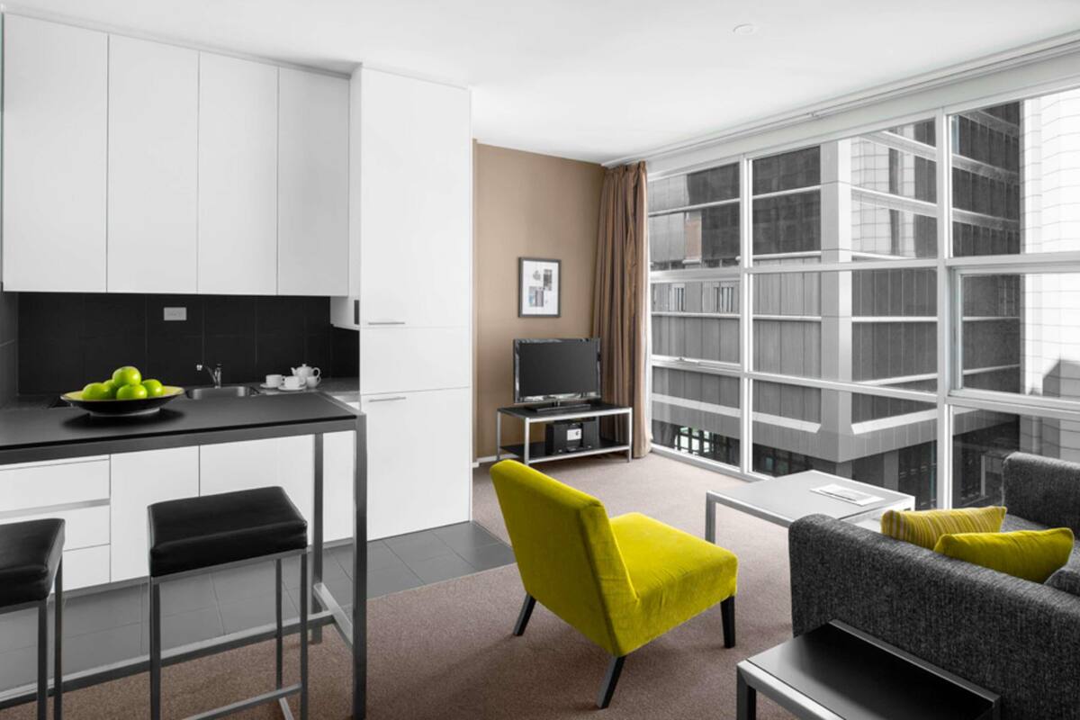 A modern living room with a sofa, a coffee table, and a TV. There is a kitchen area with cabinets and a counter.