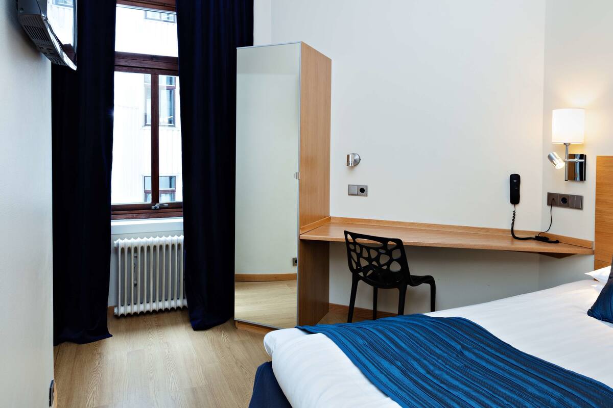 A hotel room with a bed, a desk, a chair, a radiator, a window with curtains, and a wall-mounted phone.