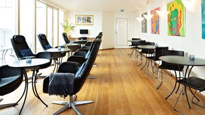 Une salle de conférence moderne, dotée de fauteuils en cuir noir, de tables rondes et d’un plancher en bois. Un grand mur vitré se trouve d’un côté, ainsi qu’une porte et un autre mur orné d’œuvres d’art colorées.
