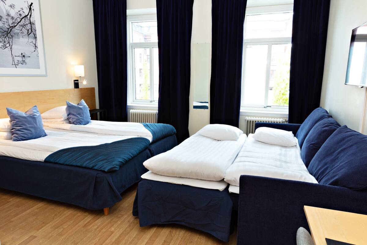 A hotel room with two beds, a sofa, a wooden floor, a window with curtains, and a wall-mounted TV.