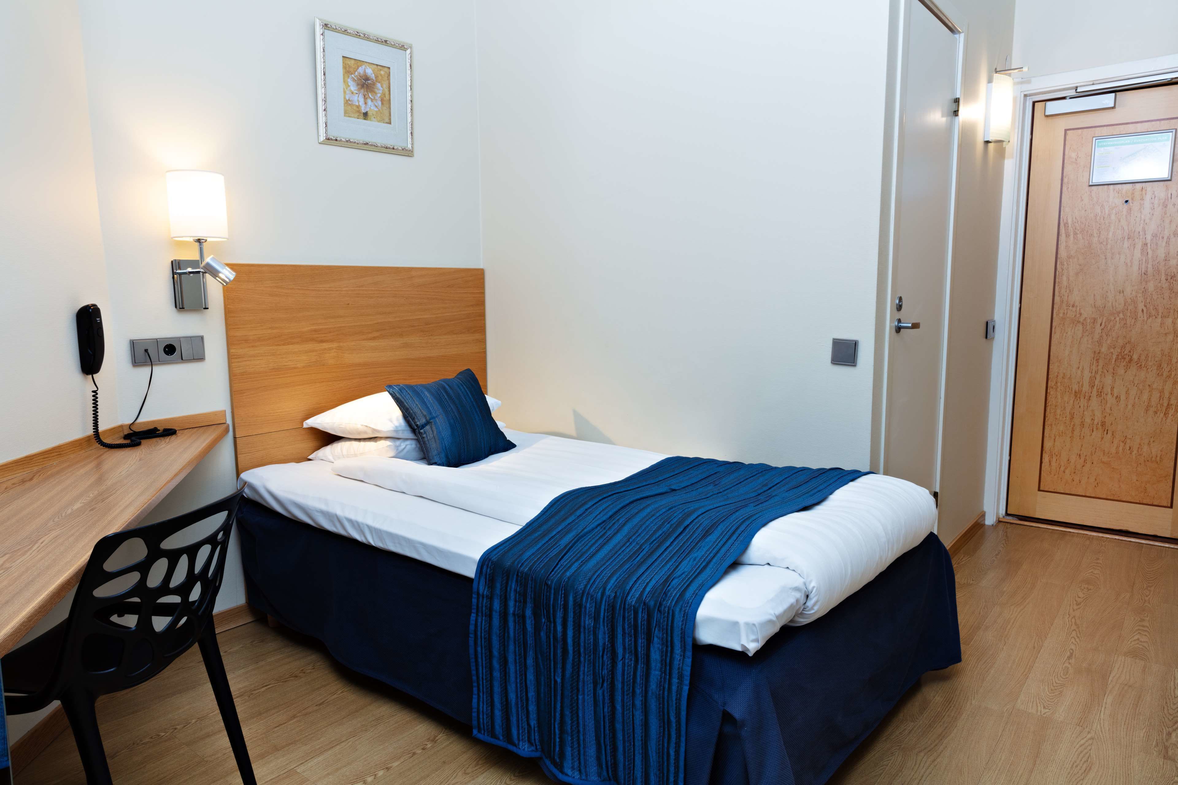 Ett hotellrum med en sänggavel i trä, ett skrivbord med en stol, en säng med ett blått överkast, en sänglampa och en dörr.