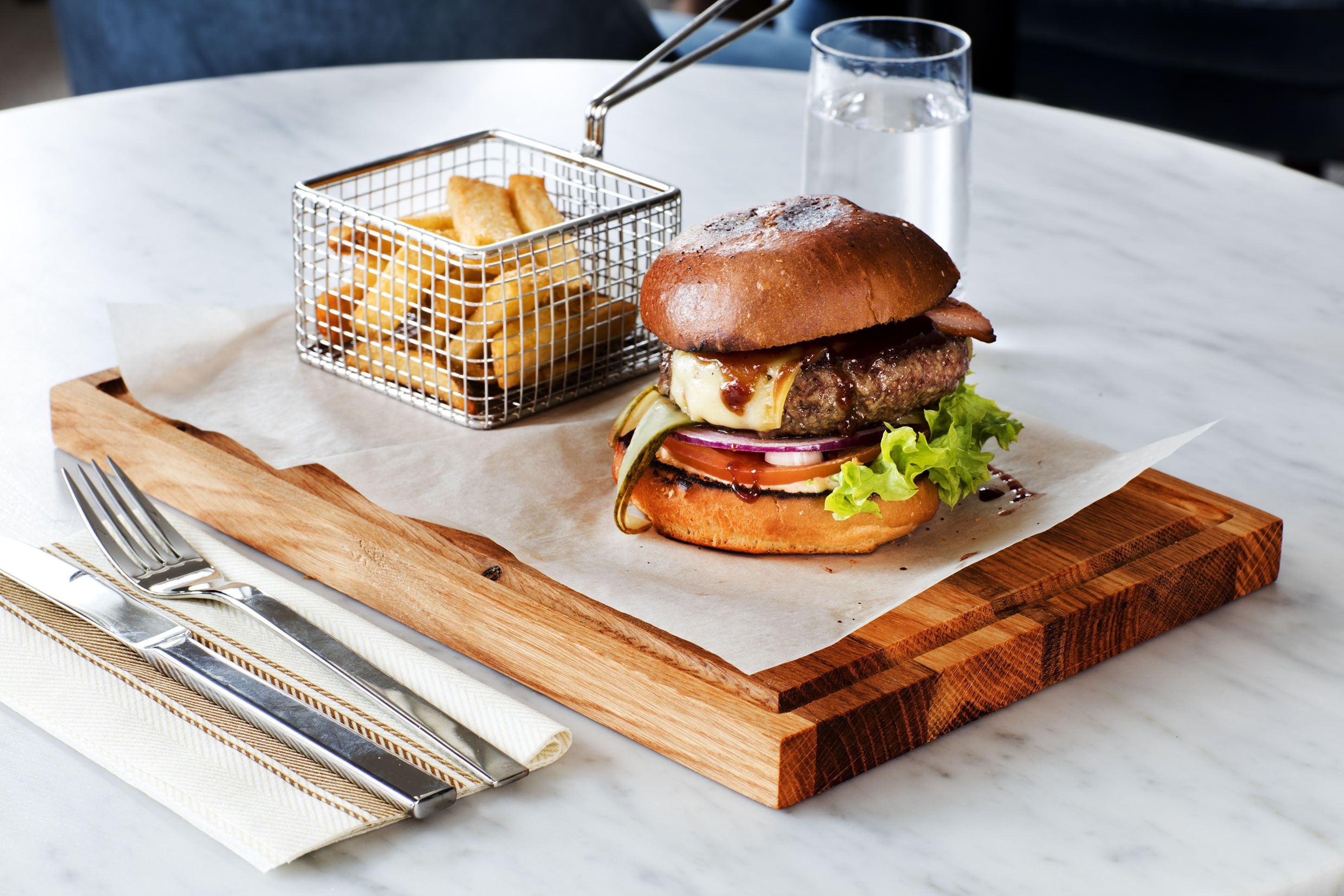 A burger with fries on a wooden board, a glass of water, and cutlery on a marble table.