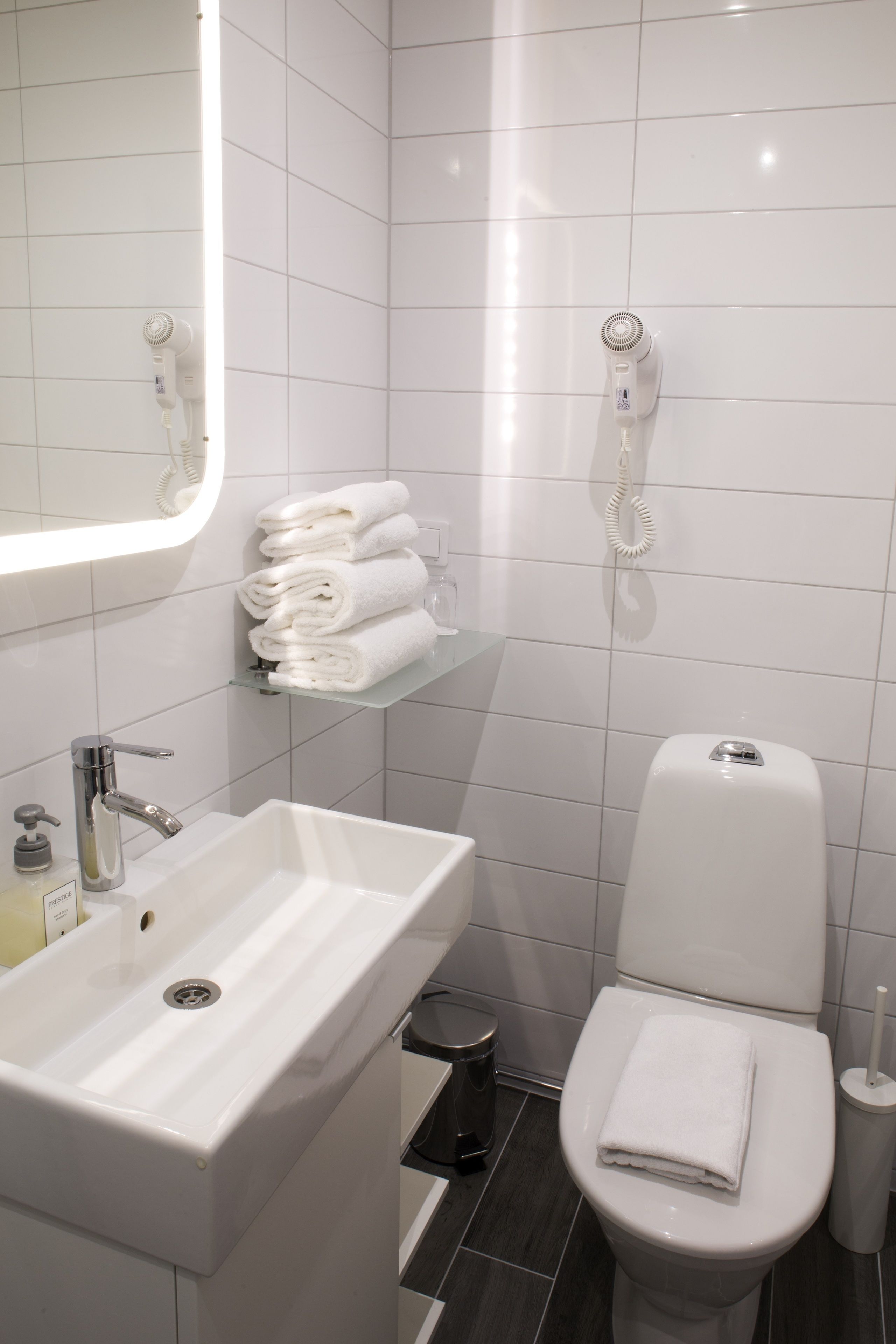 A bathroom with a white sink, a toilet, a towel rack with folded towels, and a hairdryer mounted on the wall.