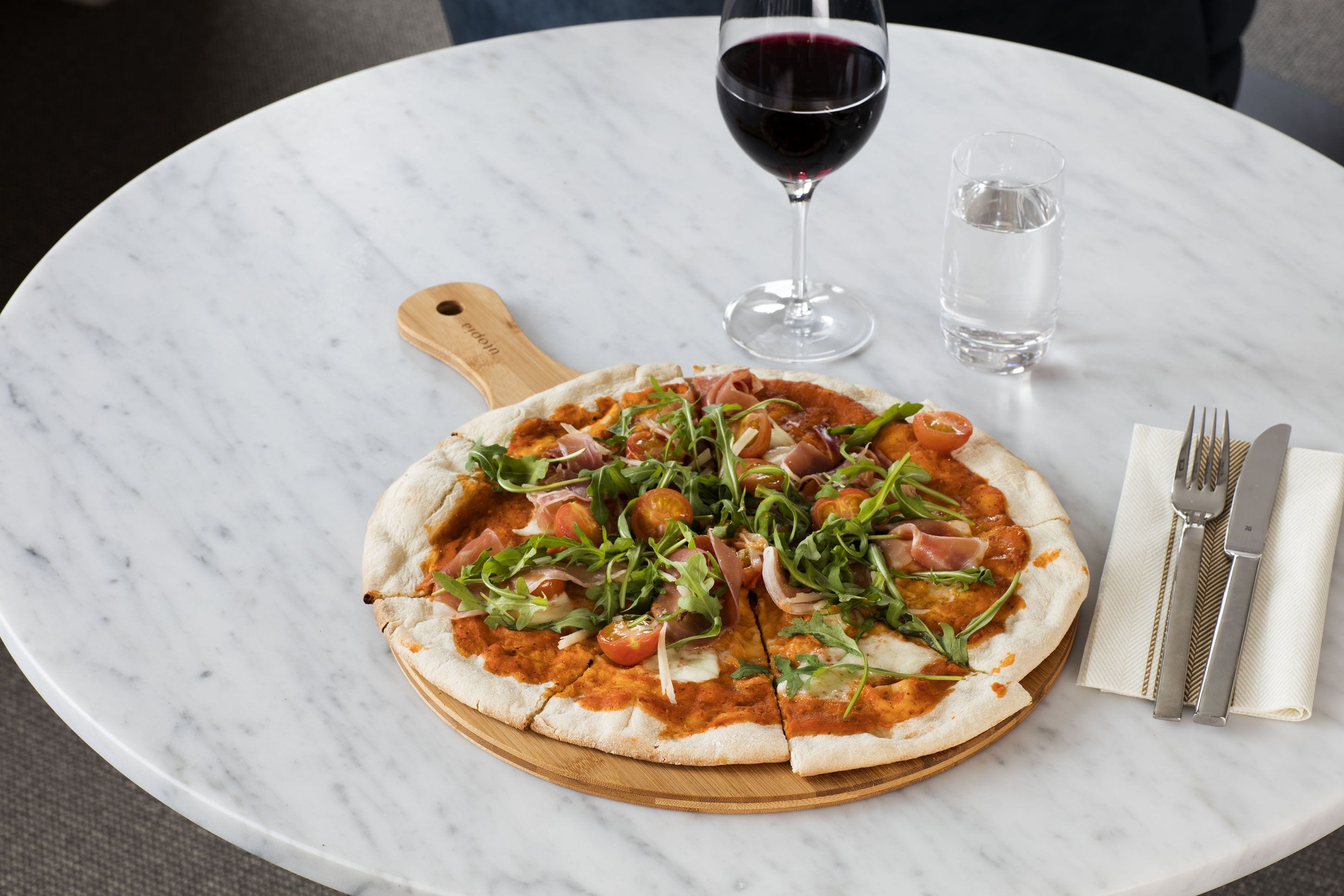 Pizza op een houten plank op een marmeren tafel, met een glas rode wijn en een glas water.
