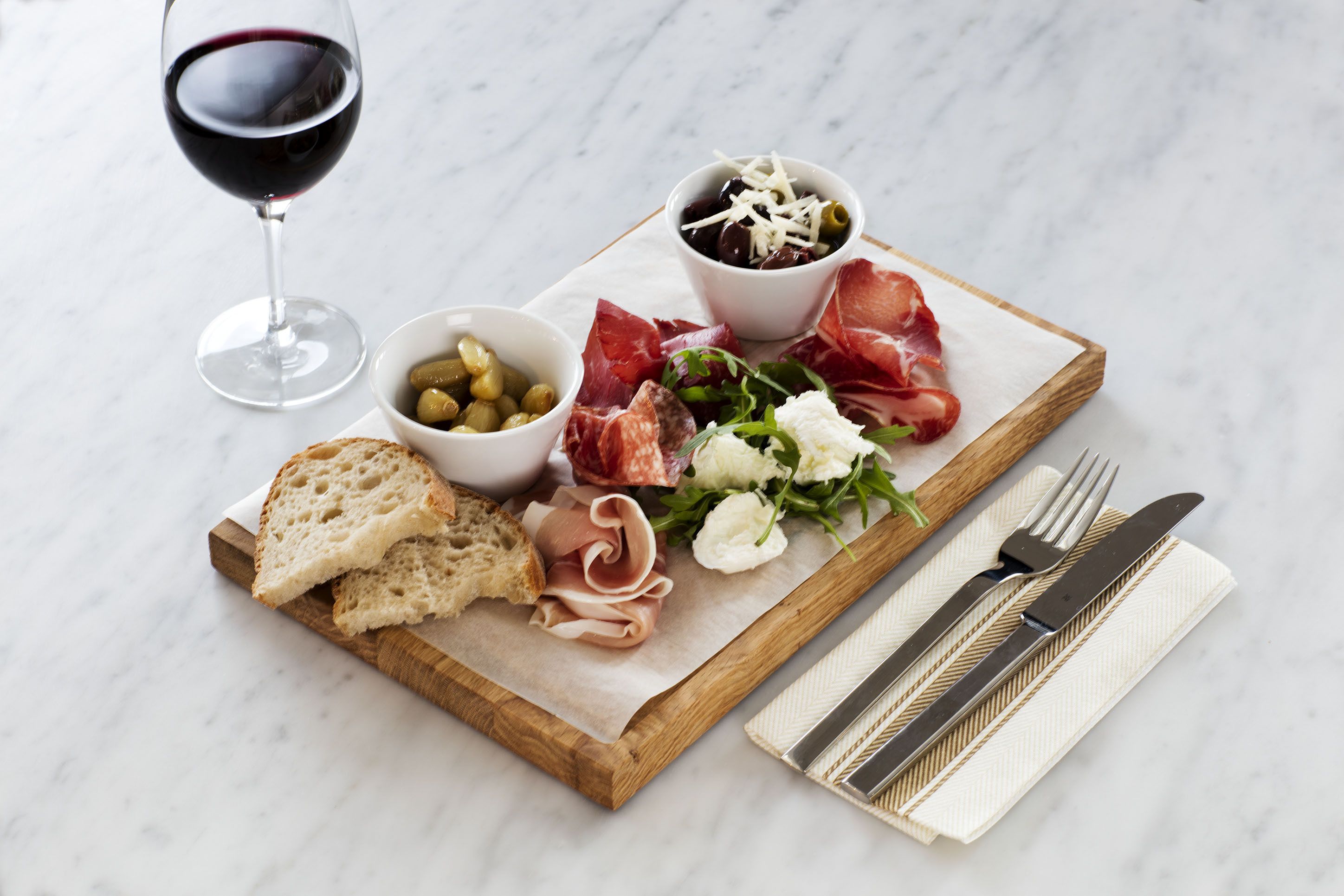 A wooden board with various food items, including bread, cheese, olives, and meats, along with a glass of red wine.