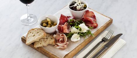 A wooden board with various food items, including bread, cheese, olives, and meats, along with a glass of red wine.