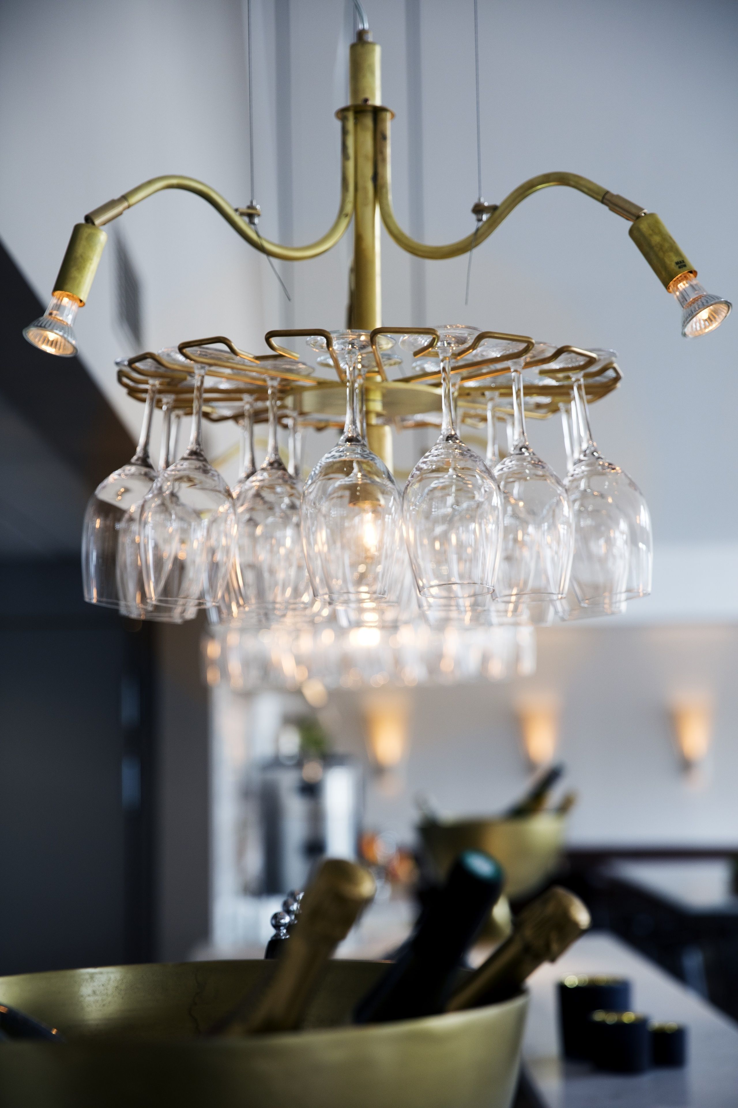 Un lampadario con diversi bicchieri da vino appesi, una ciotola di ottone con bicchieri e tappi di bottiglia, e uno sfondo sfocato che suggerisce una sala da pranzo.
