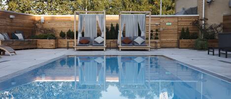 A pool area with two canopy beds, wooden deck, and a reflection in the water.