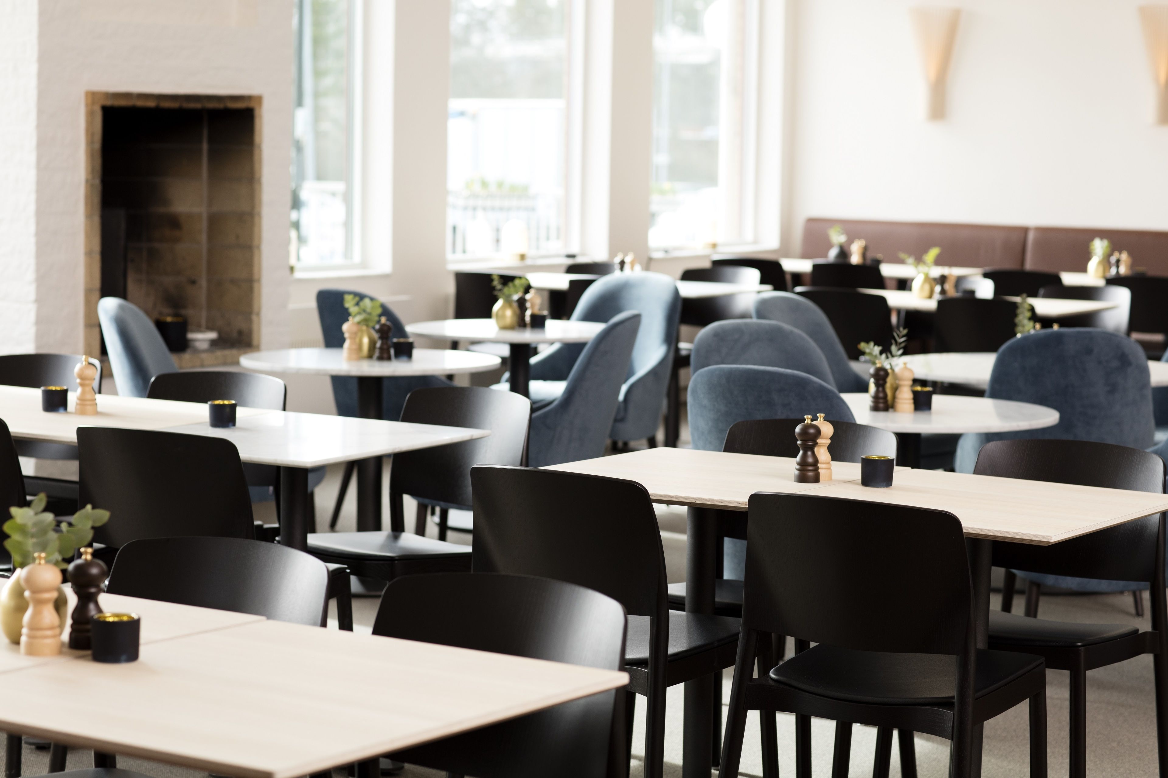 A restaurant with round tables and black chairs, a fireplace, and large windows.